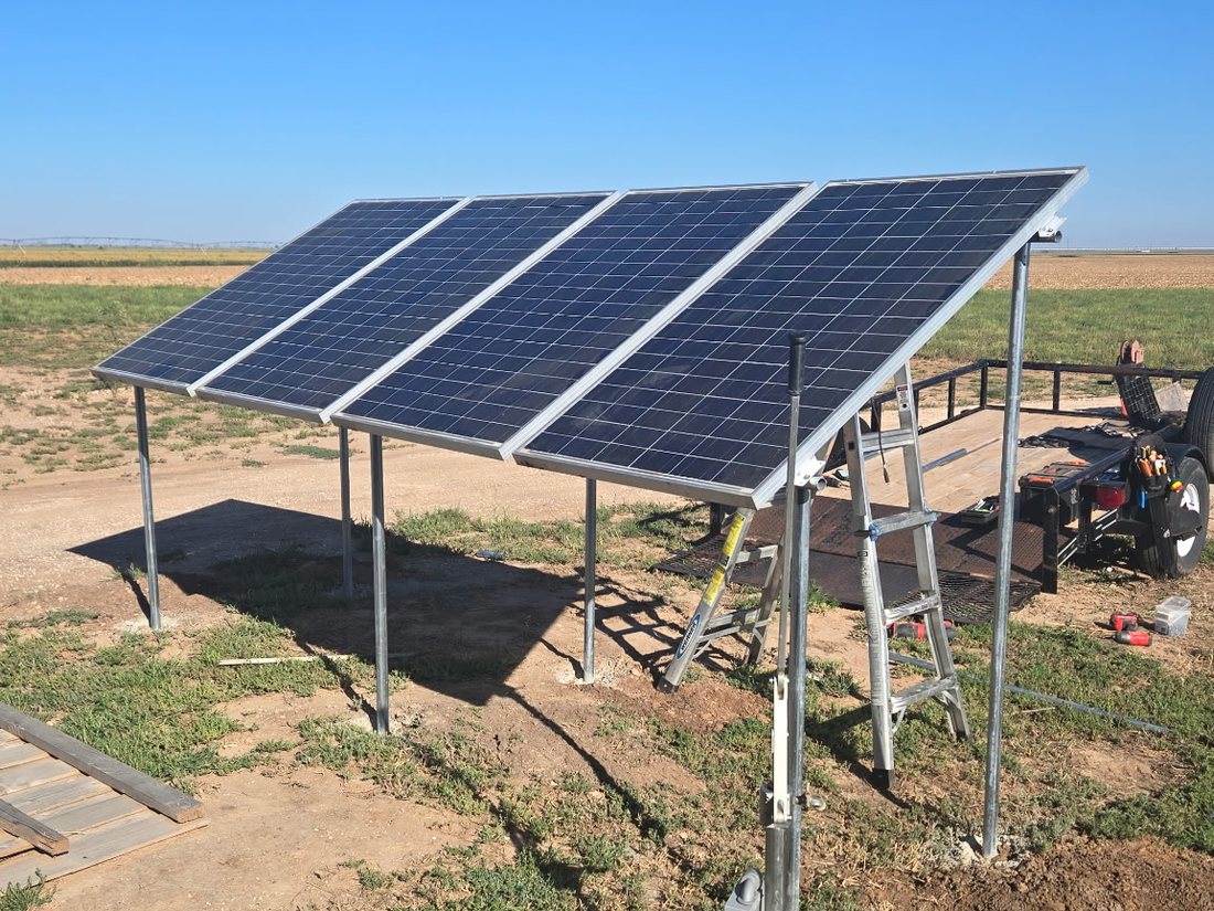 Four solar panels on a galvanized ground-mount frame beside a well head and battery box in an open Panhandle field